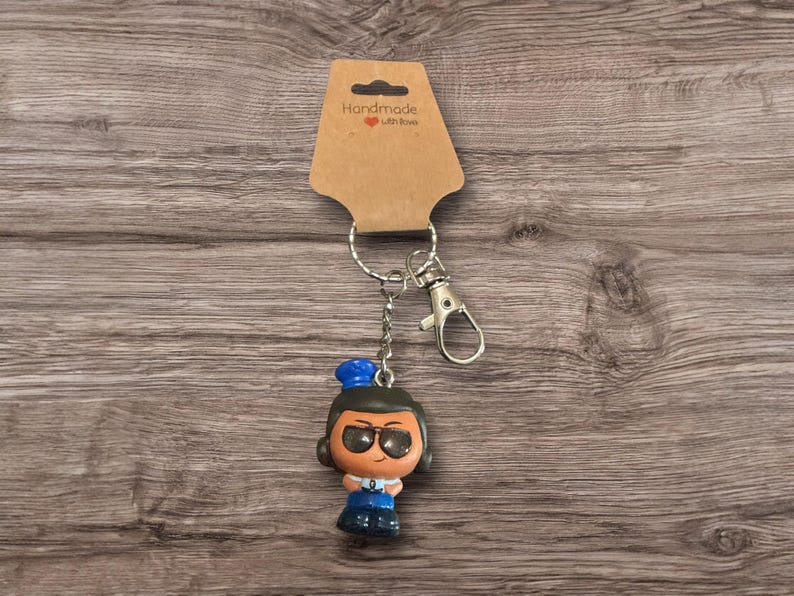Puede incluir: Un llavero con una peque&ntilde;a figura de una persona con gafas, un sombrero azul y un atuendo azul. El llavero incluye un cierre y un anillo plateados, sujetos a una etiqueta marr&oacute;n que dice "Handmade with love."