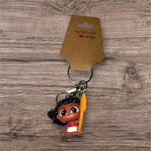 Puede incluir: Un llavero con una figura de personaje de dibujos animados con pelo oscuro, una camiseta roja y una lanza amarilla. La etiqueta marr&oacute;n dice "Handmade with love".