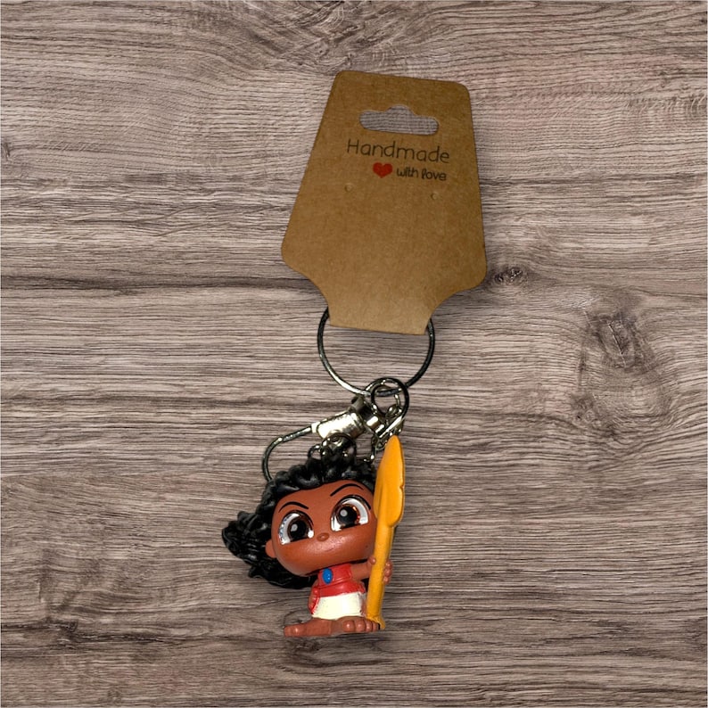 Puede incluir: Un llavero con una figura de personaje de dibujos animados con pelo oscuro, una camiseta roja y una lanza amarilla. La etiqueta marr&oacute;n dice "Handmade with love".