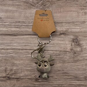May include: A grey plush reindeer keyring with large eyes and antlers. The keyring has a silver clasp and key ring, and is attached to a brown cardboard tag that reads "Handmade with Love".