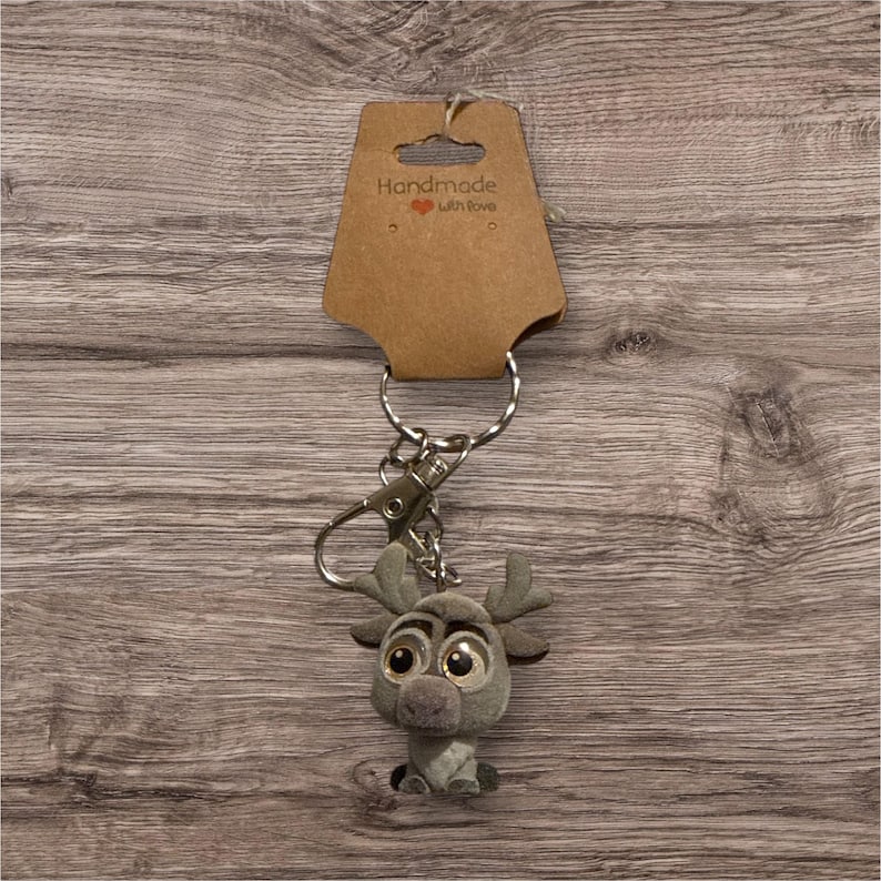 May include: A grey plush reindeer keyring with large eyes and antlers. The keyring has a silver clasp and key ring, and is attached to a brown cardboard tag that reads "Handmade with Love".