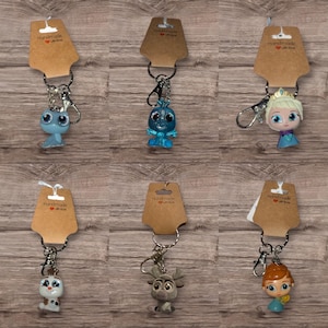 May include: Six character keyrings from a popular animated film. Each keyring is attached to a brown tag with the words "Handmade with love" printed on them. The characters include a blue dragon-like creature, a blue snowman, and a blonde princess.