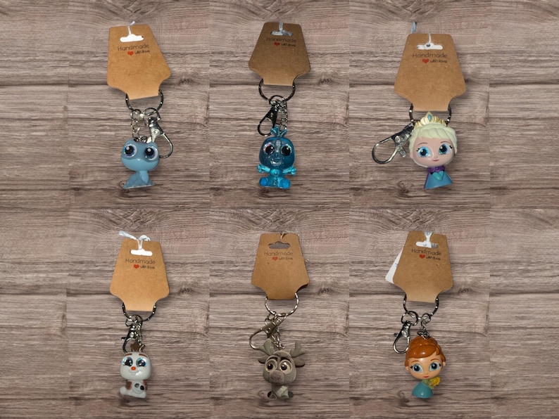 May include: Six character keyrings from a popular animated film. Each keyring is attached to a brown tag with the words "Handmade with love" printed on them. The characters include a blue dragon-like creature, a blue snowman, and a blonde princess.