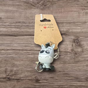 Puede incluir: Un llavero con una figura de cebra de dibujos animados en gris y blanco, con grandes ojos y una melena negra. Cuelga de una etiqueta marr&oacute;n con el texto "Handmade with Love". El fondo es una superficie de madera.