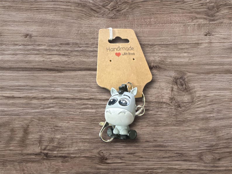 Puede incluir: Un llavero con una figura de cebra de dibujos animados en gris y blanco, con grandes ojos y una melena negra. Cuelga de una etiqueta marr&oacute;n con el texto "Handmade with Love". El fondo es una superficie de madera.