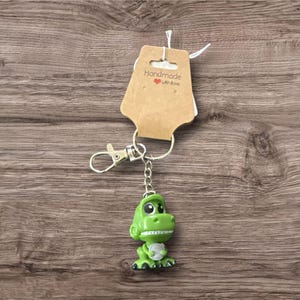 Pu&ograve; includere: Un portachiavi a forma di dinosauro verde con moschettone e catena argentati. Il dinosauro ha la pancia bianca e un'espressione amichevole. Un'etichetta marrone con la scritta "Handmade with love" &egrave; attaccata.
