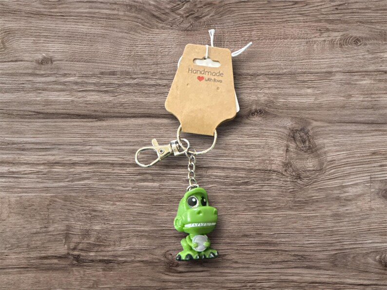 Pu&ograve; includere: Un portachiavi a forma di dinosauro verde con moschettone e catena argentati. Il dinosauro ha la pancia bianca e un'espressione amichevole. Un'etichetta marrone con la scritta "Handmade with love" &egrave; attaccata.