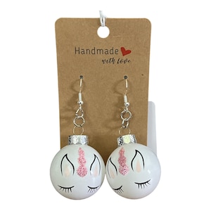 May include: A pair of white unicorn ornament earrings with pink glitter horns and silver hooks. The earrings are on a brown card with the text "Handmade with love".