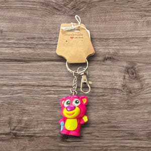 Puede incluir: Un llavero de oso rosa con detalles amarillos y un cierre plateado. El llavero est&aacute; unido a una etiqueta marr&oacute;n que dice "Handmade with love". El oso tiene ojos verdes y un accesorio azul.