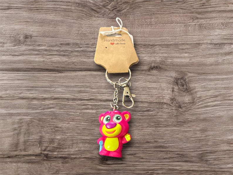 Puede incluir: Un llavero de oso rosa con detalles amarillos y un cierre plateado. El llavero est&aacute; unido a una etiqueta marr&oacute;n que dice "Handmade with love". El oso tiene ojos verdes y un accesorio azul.