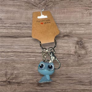 May include: A light blue keyring featuring a cartoon animal with large, glittery eyes. The keyring is attached to a silver metal ring and clasp, and is displayed with a brown tag that reads "Handmade with Love".