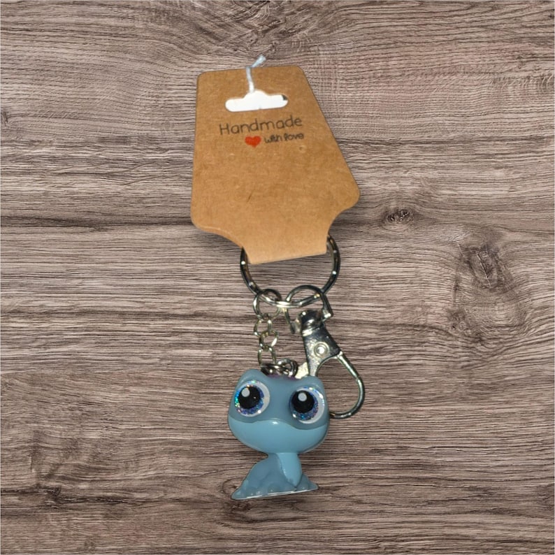 May include: A light blue keyring featuring a cartoon animal with large, glittery eyes. The keyring is attached to a silver metal ring and clasp, and is displayed with a brown tag that reads "Handmade with Love".