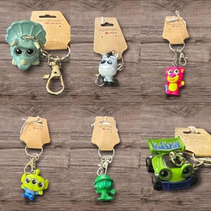 Puede incluir: Colecci&oacute;n de llaveros de personajes, que incluyen un dinosaurio verde, un burro gris, un oso rosa, un extraterrestre verde, un extraterrestre verde y un coche verde. Cada llavero est&aacute; unido a una etiqueta marr&oacute;n con las palabras "Handmade with love".