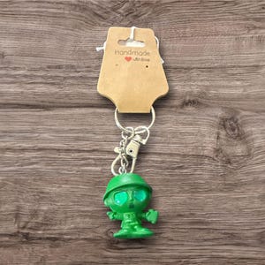 Puede incluir: Un llavero de soldado de juguete verde con un llavero y un cierre plateados. El soldado lleva un casco. Una etiqueta marr&oacute;n con la inscripci&oacute;n "Handmade with love" est&aacute; adjunta.