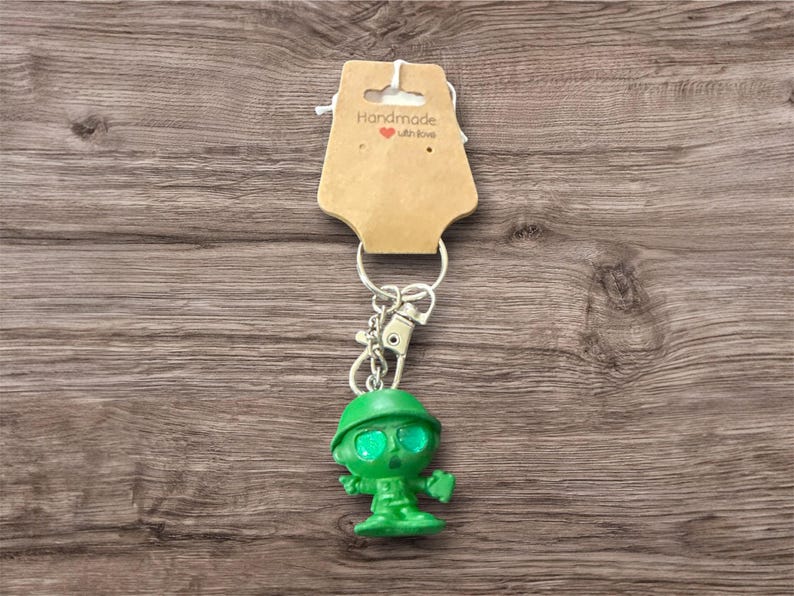 Puede incluir: Un llavero de soldado de juguete verde con un llavero y un cierre plateados. El soldado lleva un casco. Una etiqueta marr&oacute;n con la inscripci&oacute;n "Handmade with love" est&aacute; adjunta.