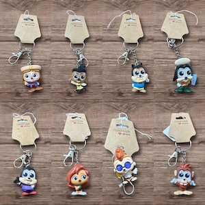 May include: A collection of handmade character keychains. Each keychain features a small, colorful figurine attached to a silver-tone metal keyring and clasp. The figurines depict various cartoon characters. Each keychain has a brown tag with the words "Handmade with love".