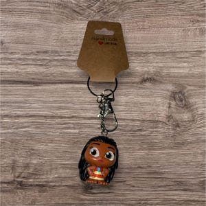 May include: A keychain featuring a small, cartoon-style figure with black hair and a colorful outfit. The figure is attached to a silver-colored chain and clasp, with a brown tag that reads "Handmade with love."