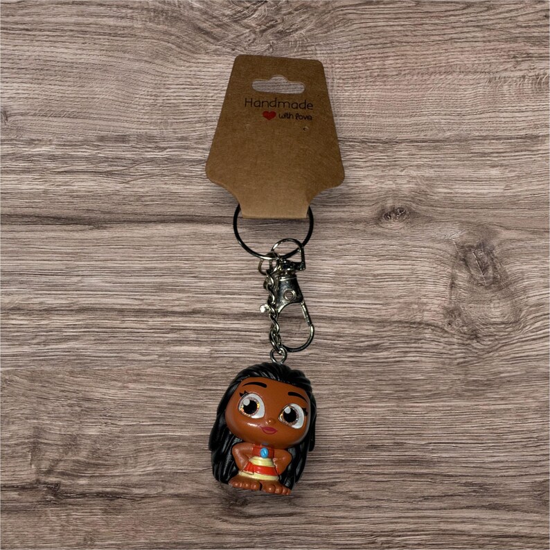 May include: A keychain featuring a small, cartoon-style figure with black hair and a colorful outfit. The figure is attached to a silver-colored chain and clasp, with a brown tag that reads "Handmade with love."