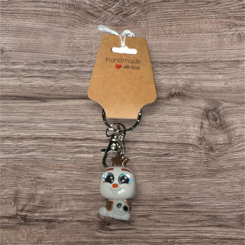 May include: A keyring with a small, white figurine with large, glittery blue eyes, a carrot nose, and small brown antlers. The figurine is attached to a silver keyring and a brown tag that reads "Handmade with love."