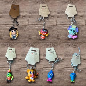 May include: Collection of handmade keychains featuring various cartoon characters. Each keychain has a silver-tone chain and clasp, attached to a small, colorful figurine. The figurines include a frog, a bear, a blue bird, and other whimsical designs.
