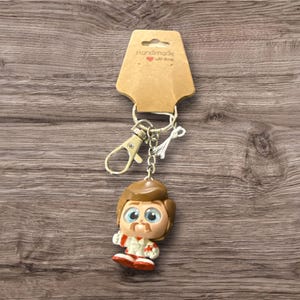 May include: A keychain featuring a cartoon character with brown hair, a mustache, and a red and white outfit. The keychain is attached to a metal clasp and a brown cardboard tag that reads "Handmade with love".