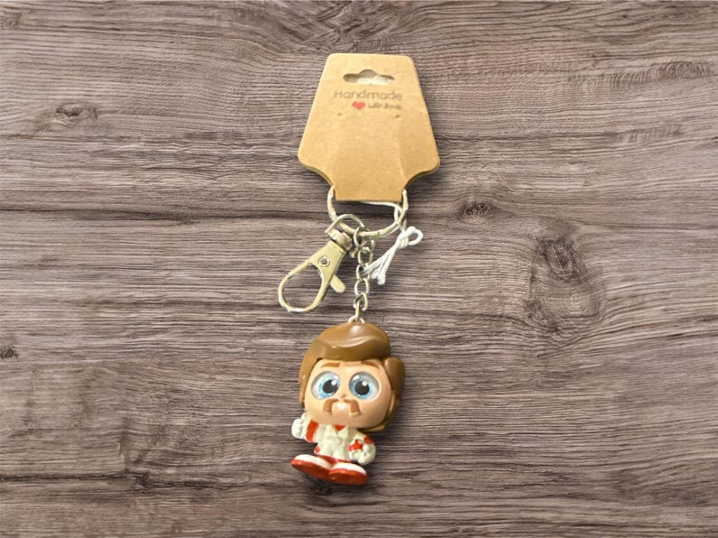 May include: A keychain featuring a cartoon character with brown hair, a mustache, and a red and white outfit. The keychain is attached to a metal clasp and a brown cardboard tag that reads "Handmade with love".