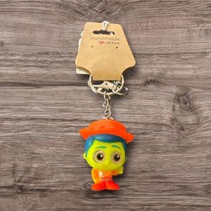 Puede incluir: Un llavero colorido con un personaje de dibujos animados con un sombrero naranja, pelo azul y una cara amarilla. El llavero est&aacute; unido a una cadena plateada y una etiqueta marr&oacute;n con el texto "Handmade with love".