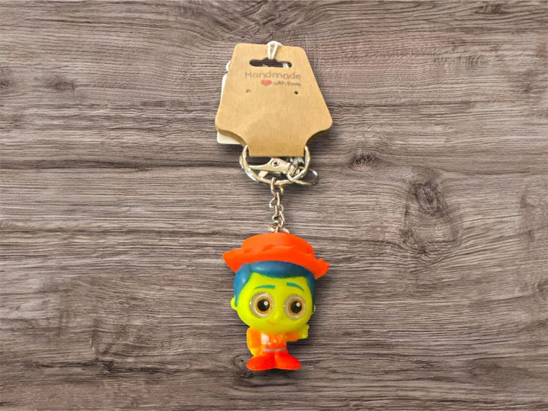 Puede incluir: Un llavero colorido con un personaje de dibujos animados con un sombrero naranja, pelo azul y una cara amarilla. El llavero est&aacute; unido a una cadena plateada y una etiqueta marr&oacute;n con el texto "Handmade with love".