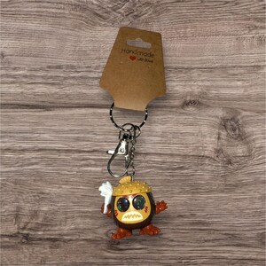 May include: A handmade keychain featuring a cartoon character with a brown body, orange feet and hands, and a yellow hat. The character has large, glittery eyes and a white object in its hand. The keychain includes a metal clasp and a ring, with a brown tag that reads "Handmade with love."
