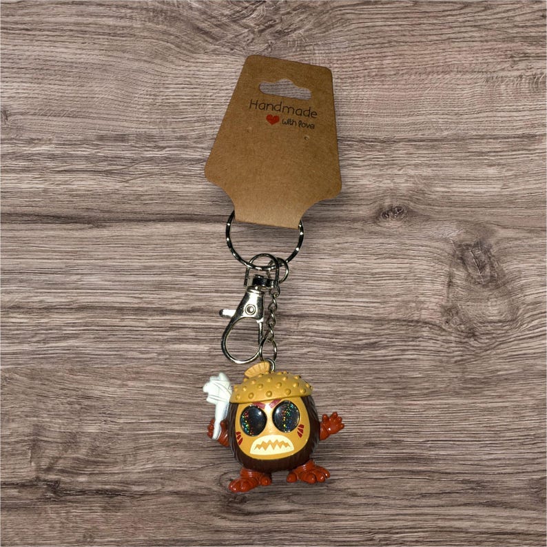 May include: A handmade keychain featuring a cartoon character with a brown body, orange feet and hands, and a yellow hat. The character has large, glittery eyes and a white object in its hand. The keychain includes a metal clasp and a ring, with a brown tag that reads "Handmade with love."