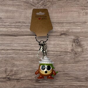 May include: A handmade keychain featuring a small, cartoonish figure with a brown body, large eyes, and a white hat. The figure has a green headband and a small green stick. The keychain includes a metal clasp and a ring, attached to a brown tag that reads "Handmade with love."