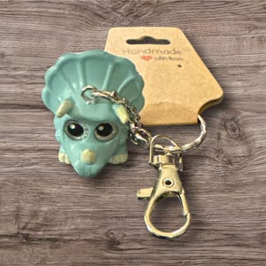 Puede incluir: Un llavero de triceratops turquesa con ojos grandes y brillantes y una cadena y cierre plateados. El llavero est&aacute; unido a una etiqueta marr&oacute;n que dice "Handmade with love."