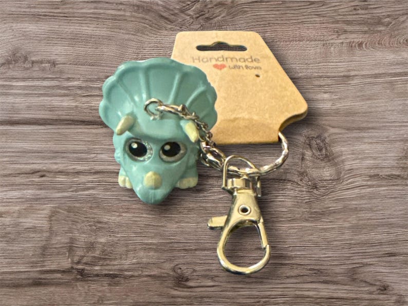 Puede incluir: Un llavero de triceratops turquesa con ojos grandes y brillantes y una cadena y cierre plateados. El llavero est&aacute; unido a una etiqueta marr&oacute;n que dice "Handmade with love."