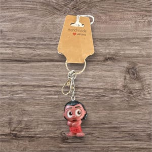 Puede incluir: Un llavero hecho a mano con un personaje de dibujos animados con cabello oscuro y ropa roja. El llavero tiene un cierre y un llavero de color plateado, y est&aacute; sujeto a una etiqueta marr&oacute;n que dice "Handmade with love". El fondo es una superficie de madera.
