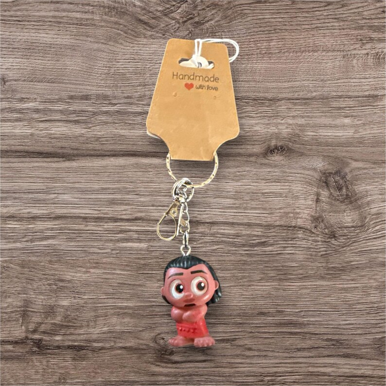 Puede incluir: Un llavero hecho a mano con un personaje de dibujos animados con cabello oscuro y ropa roja. El llavero tiene un cierre y un llavero de color plateado, y est&aacute; sujeto a una etiqueta marr&oacute;n que dice "Handmade with love". El fondo es una superficie de madera.