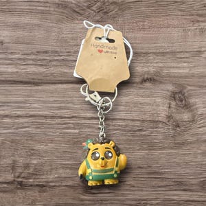 May include: A handmade keychain featuring a yellow cartoon character with green overalls and a small red bow. The keychain has a silver chain and clasp, and a brown tag that reads "Handmade with love."