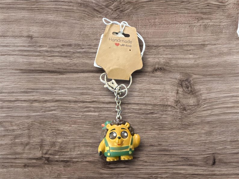 May include: A handmade keychain featuring a yellow cartoon character with green overalls and a small red bow. The keychain has a silver chain and clasp, and a brown tag that reads "Handmade with love."