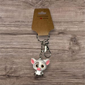 May include: A keychain featuring a small, cartoon pig with large, glittery eyes, pink ears, and a pink snout. The pig is white and gray, attached to a silver chain and clasp. The tag reads "Handmade with love."