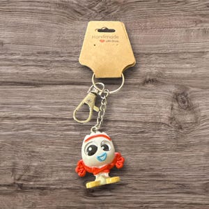 Pu&ograve; includere: Un portachiavi con un personaggio dei cartoni animati. Il personaggio ha una testa bianca, occhi blu e braccia rosse. Il portachiavi ha un moschettone e una catena argentati ed &egrave; attaccato a un'etichetta marrone con "Handmade with love."
