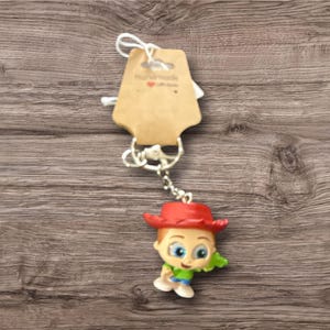 May include: A keychain featuring a small, colorful figurine of a cartoon character wearing a red hat, green shirt, and blue pants. The keychain has a silver chain and a brown tag with the text "Handmade with love".