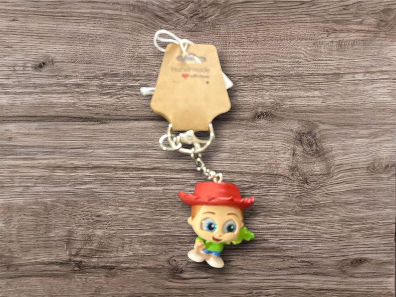 May include: A keychain featuring a small, colorful figurine of a cartoon character wearing a red hat, green shirt, and blue pants. The keychain has a silver chain and a brown tag with the text "Handmade with love".