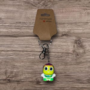 Puede incluir: Un llavero hecho a mano con una peque&ntilde;a y colorida figura alien&iacute;gena con ojos grandes, cabeza morada y brazos y piernas blancos. El llavero incluye un anillo de metal, un cierre y una etiqueta marr&oacute;n que dice "Handmade with love".