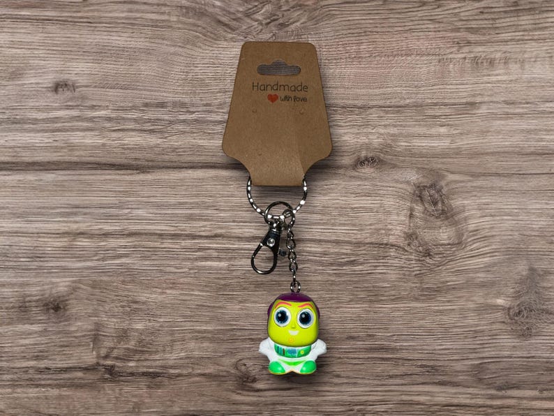 Puede incluir: Un llavero hecho a mano con una peque&ntilde;a y colorida figura alien&iacute;gena con ojos grandes, cabeza morada y brazos y piernas blancos. El llavero incluye un anillo de metal, un cierre y una etiqueta marr&oacute;n que dice "Handmade with love".
