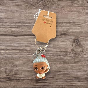 Puede incluir: Un llavero con una peque&ntilde;a figura de una anciana con pelo gris y una flor roja. La figura est&aacute; unida a un llavero plateado y una etiqueta marr&oacute;n que dice "Handmade with love."