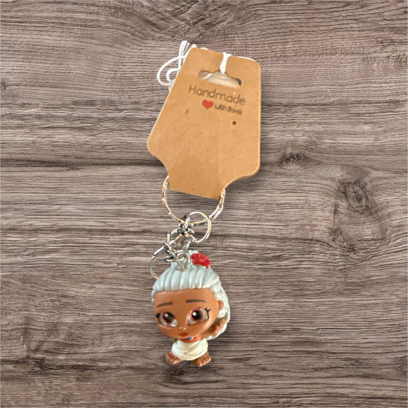 Puede incluir: Un llavero con una peque&ntilde;a figura de una anciana con pelo gris y una flor roja. La figura est&aacute; unida a un llavero plateado y una etiqueta marr&oacute;n que dice "Handmade with love."