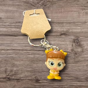 May include: A keychain featuring a small, doll-like figure with brown hair, yellow accents, and large green eyes. The figure is attached to a silver chain and a cardboard tag. The background is a wooden surface.