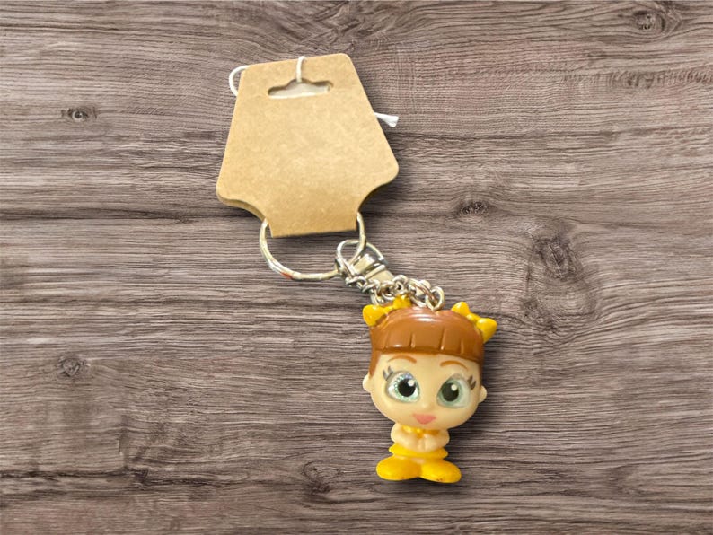 May include: A keychain featuring a small, doll-like figure with brown hair, yellow accents, and large green eyes. The figure is attached to a silver chain and a cardboard tag. The background is a wooden surface.