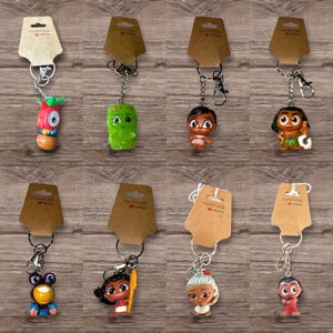 Puede incluir: Una colecci&oacute;n de llaveros de personajes con etiquetas de cart&oacute;n marr&oacute;n. Los llaveros presentan varias figuras de dibujos animados coloridas, unidas a llaveros plateados. El fondo es una superficie de madera.