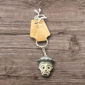 May include: A keychain featuring a small, detailed figurine of a character with a hat and a neutral color palette. The keychain is attached to a metal ring and a small tag. The background is a wooden surface.