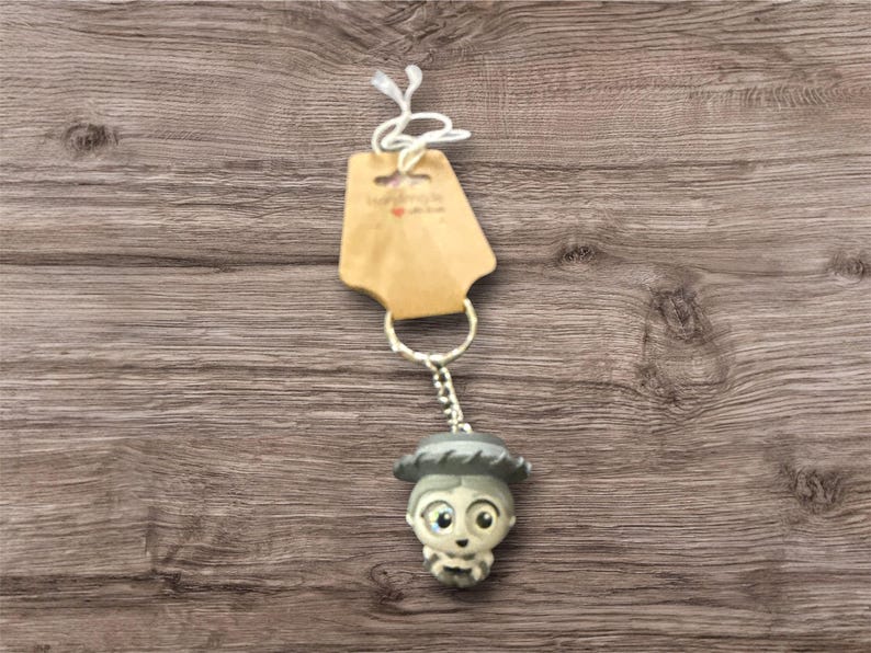 May include: A keychain featuring a small, detailed figurine of a character with a hat and a neutral color palette. The keychain is attached to a metal ring and a small tag. The background is a wooden surface.